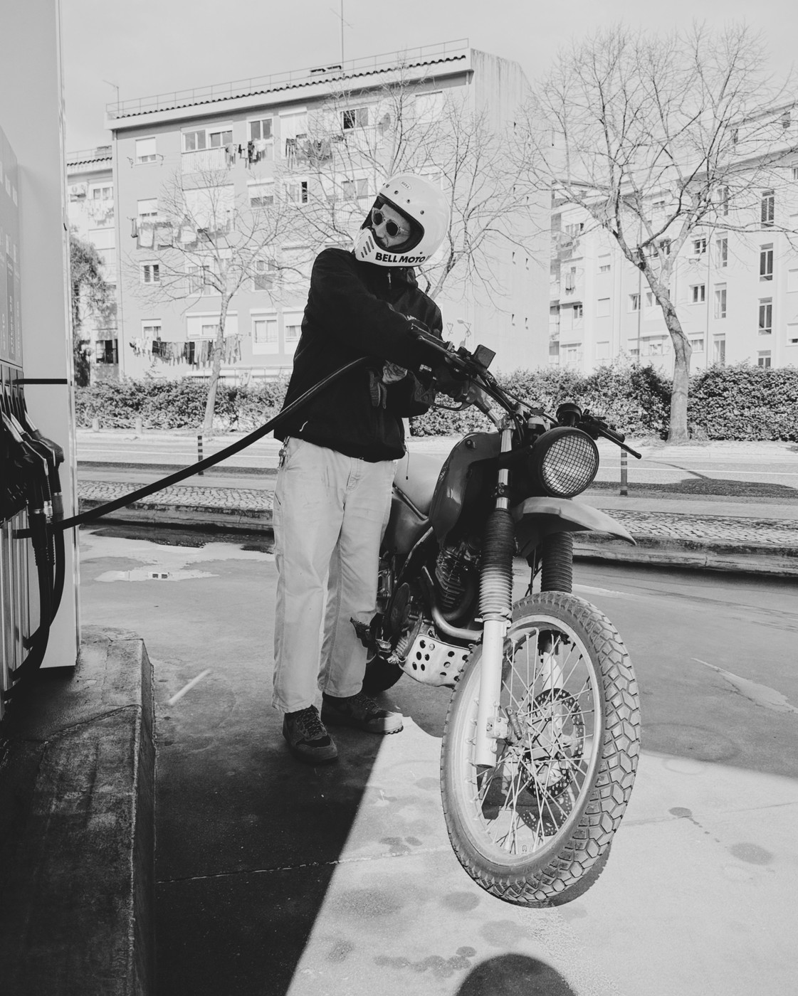 A black-and-white photo of a person in a white Bell Moto helmet refueling a dual-sport motorcycle at a gas station. Behind them are bare trees and a residential building.