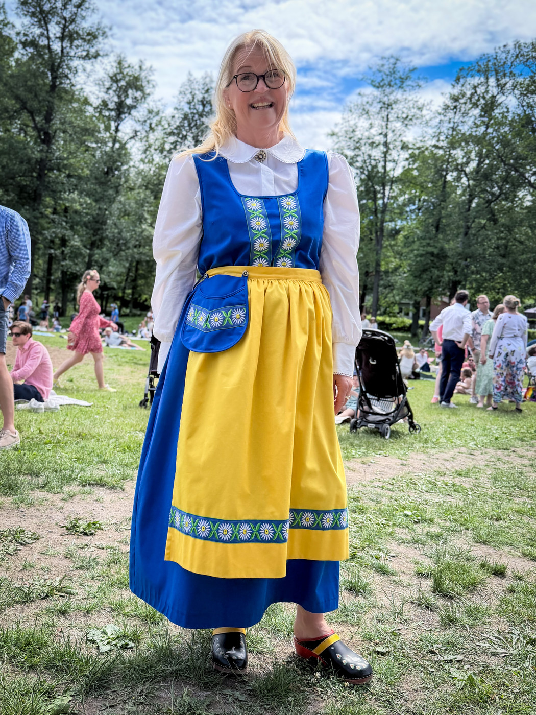 A smiling woman in a Swedish folk costume, with a blue dress, white blouse, yellow apron with flower trim and wooden clogs, standing on a grassy field with people picnicking behind her.