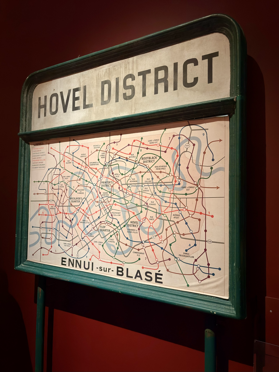 Freestanding sign titled ‘Hovel District’ displaying a dense, colour-coded transit-style map with district names and routes, framed in green against a dark red background.