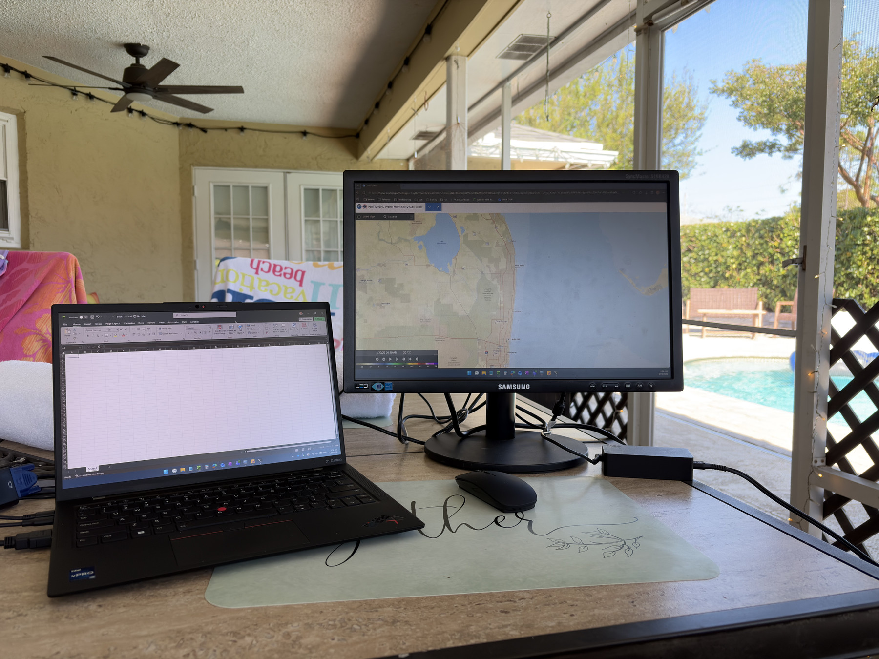 A laptop and monitor on a table near a pool, with a map on the screen and a fan overhead.