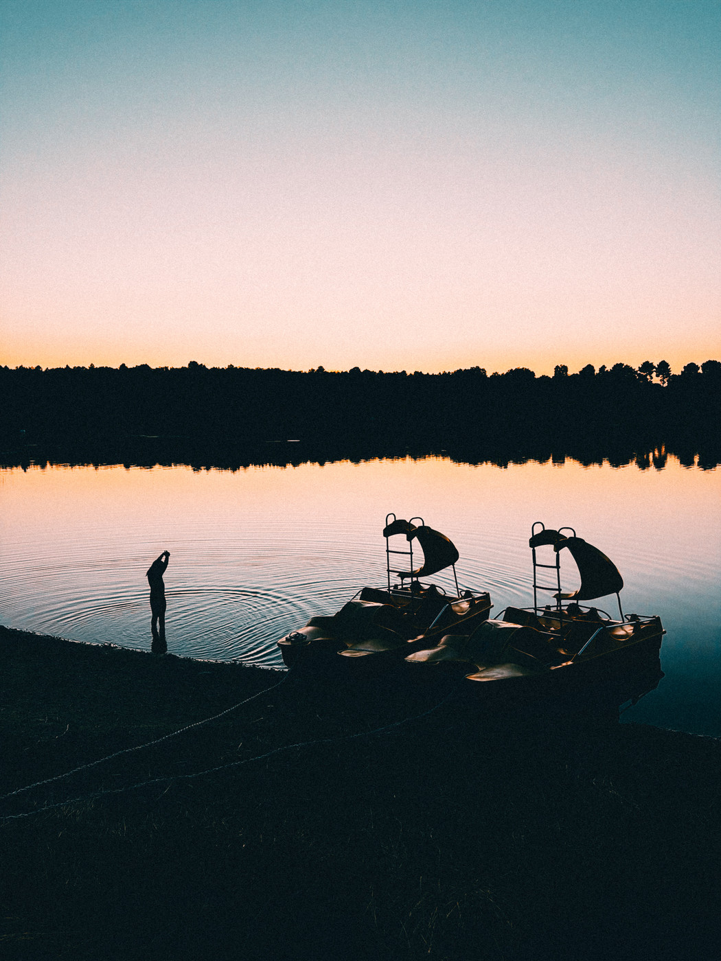 Silhouette of a person standing in calm water near two pedal boats at sunset, with a tree-lined horizon and clear sky reflected in the lake.