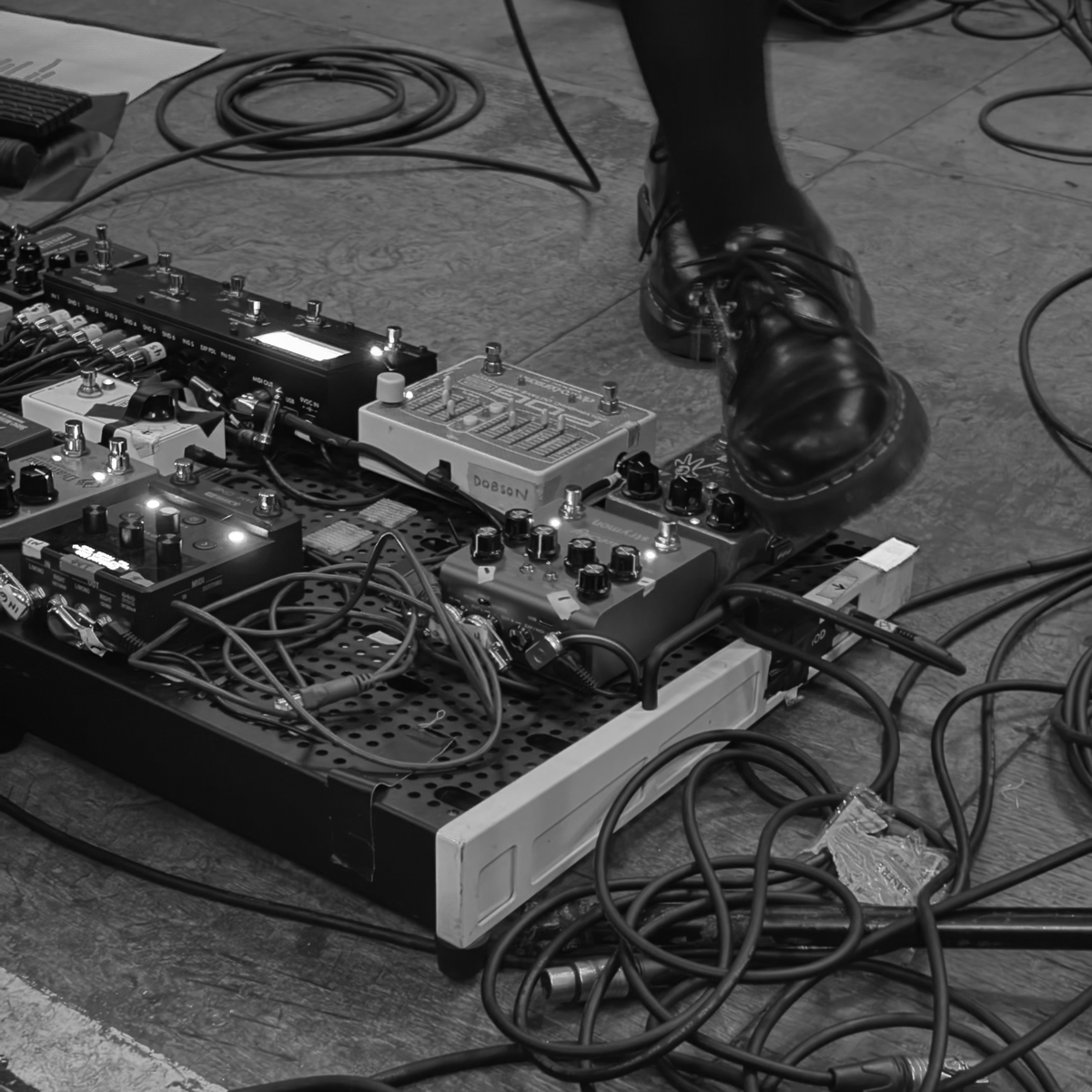 A black-and-white photo showing a musician’s foot stepping near a pedalboard filled with multiple guitar effects pedals and coiled cables on a stage floor.