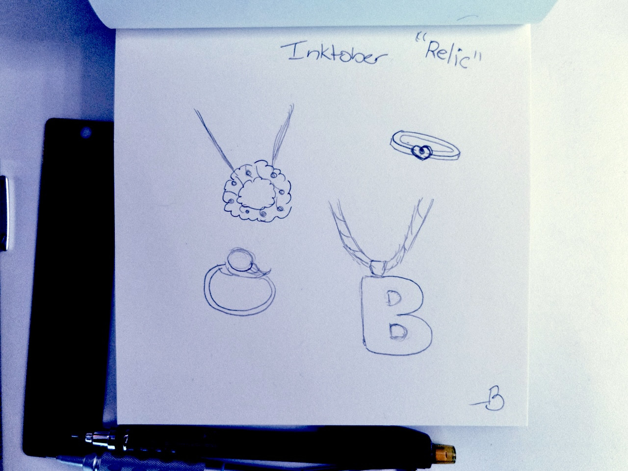 A snapshot of a square notebook with blank paper, and drawn on the page are a few pieces of jewelry for Inktober’s theme, “relic”.