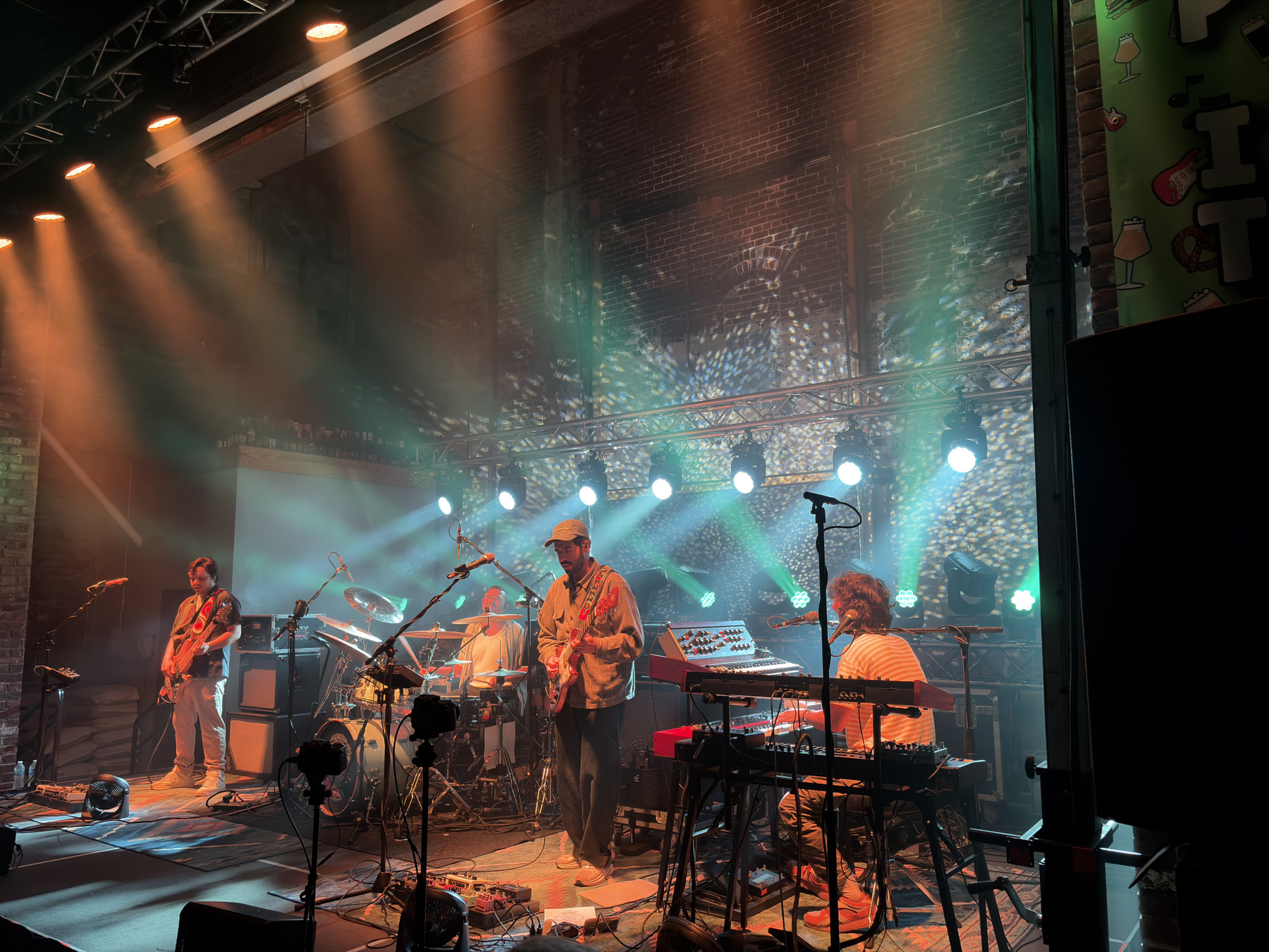 A band performing on stage with colorful lights, instruments, and a brick wall backdrop, creating a lively concert atmosphere.