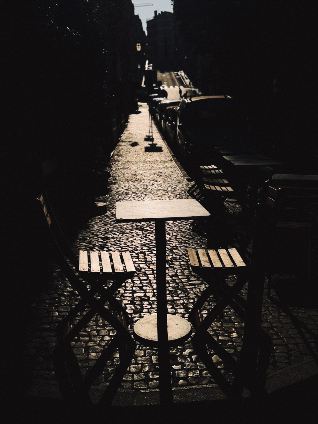 Silhouetted outdoor café table with two chairs on a cobblestone street, with cars parked along the side and a distant view of the street uphill.