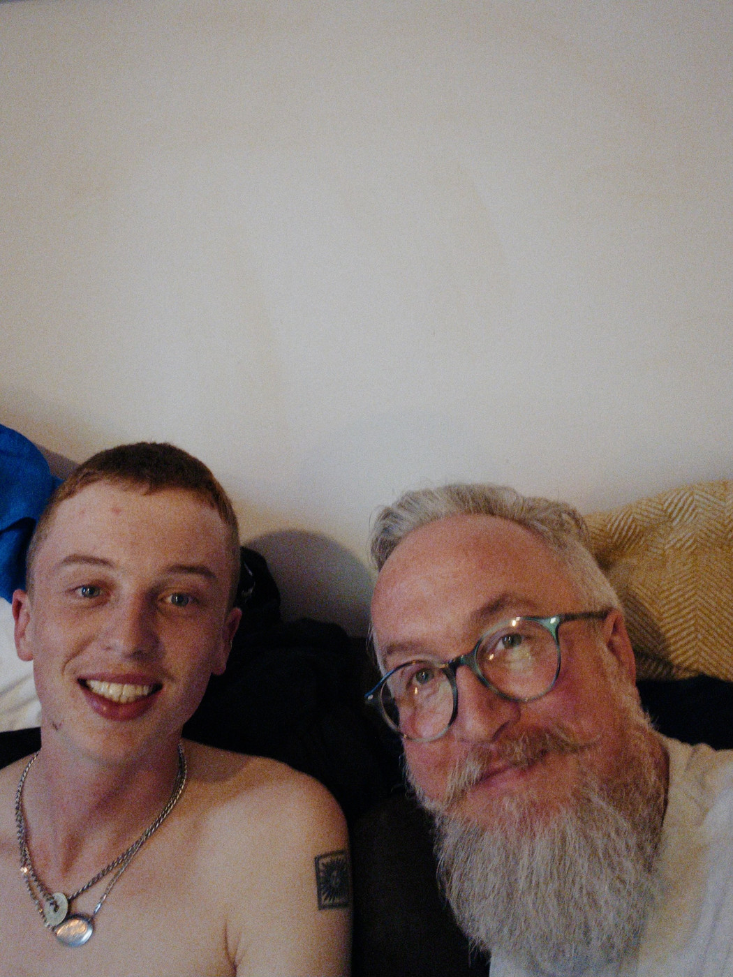 Indoor selfie of two people sitting close together and smiling toward the camera. One person wears glasses and has a long grey beard, while the other wears layered necklaces and has a small tattoo on their upper arm. Soft, warm lighting and a plain wall create a relaxed, candid atmosphere.