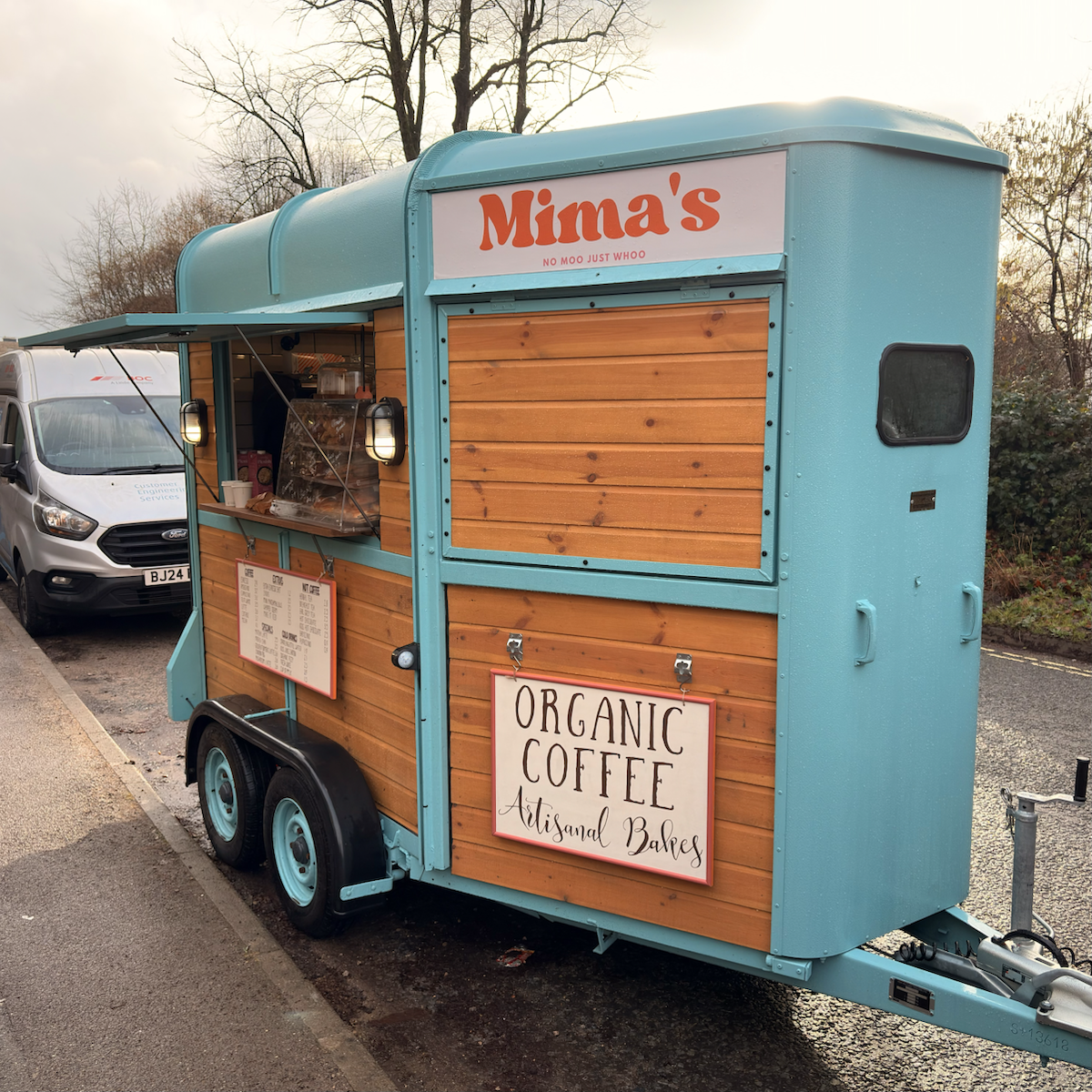 Turquoise and wood-paneled mobile coffee trailer parked roadside, with a serving hatch open, a sign reading Mima’s, and another sign advertising organic coffee and artisanal bakes.