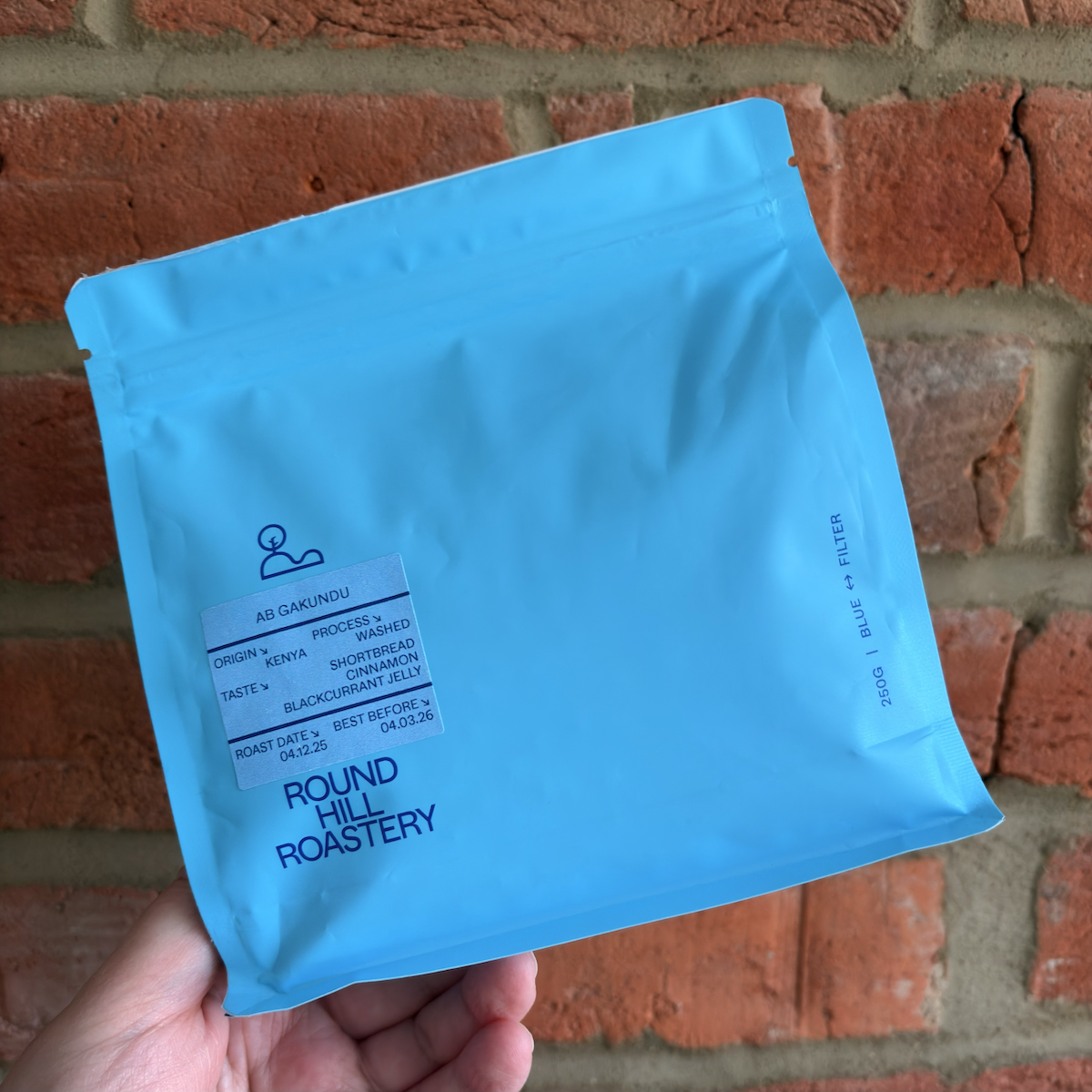 A bright blue coffee bag from Round Hill Roastery held in one hand against a red brick wall, with a label showing Kenyan washed coffee “AB Gakundu” and tasting notes of shortbread, cinnamon, and blackcurrant jelly.
