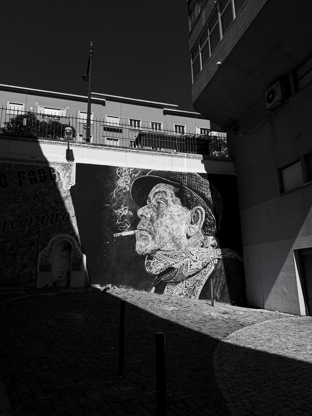 Black and white photo of a street mural depicting a man in a hat smoking a cigarette, with smoke rising, on a shadowed cobblestone alley bordered by buildings.