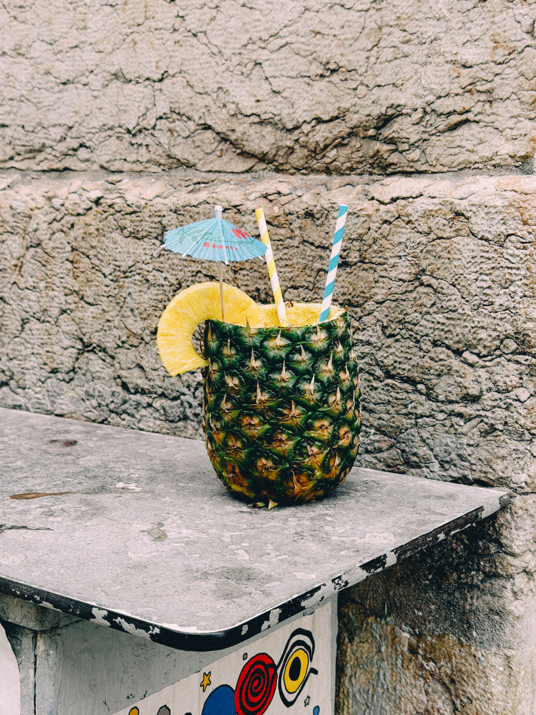 A tropical drink served in a hollowed pineapple with a lemon slice, two striped straws, and a small blue paper umbrella, placed on a weathered stone ledge against a textured wall.