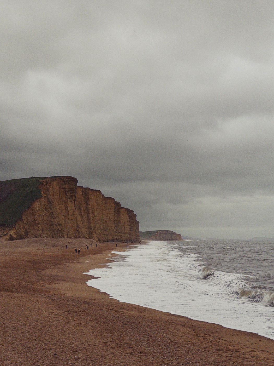 A sandy beach with a few people walking along it. The beach is bordered by tall, steep cliffs on the left. The sky is overcast and grey, and the sea is choppy with white waves crashing onto the shore. The overall mood of the image is calm and serene, with a hint of melancholy due to the grey sky.
