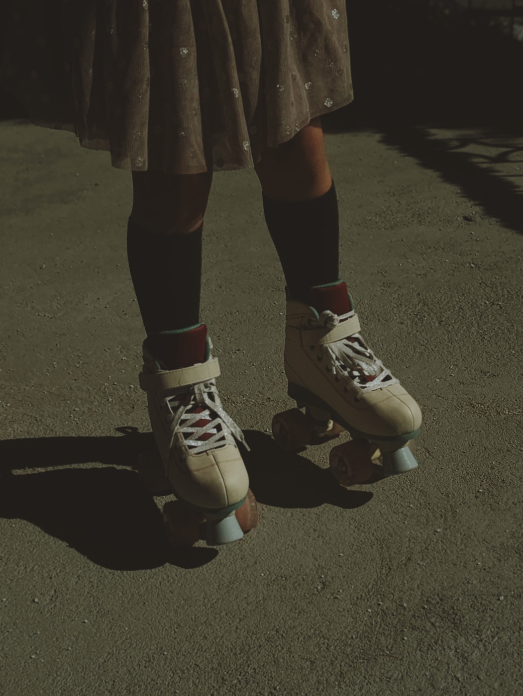 Person wearing vintage white roller skates with black knee-high socks and a brown skirt standing on a sunlit concrete surface.