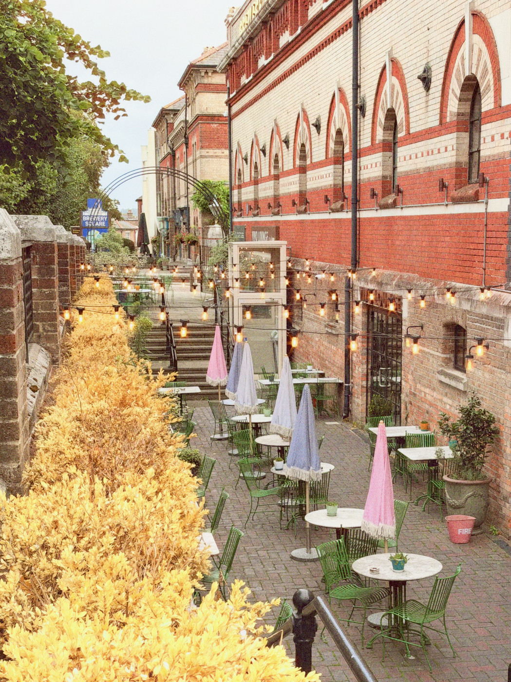 Courtyard cafe seating with green chairs, pastel striped umbrellas, and string lights beside a red-brick building, “Brewery Square” sign visible.