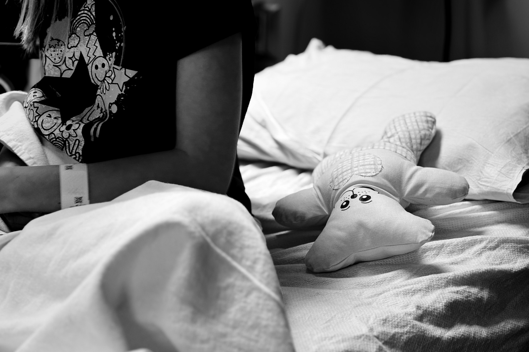 Black-and-white photo of a person sitting on a bed with a blanket over their lap, wearing a graphic T-shirt and a wristband. A small plush toy with a smiling face lies on the bed beside them.