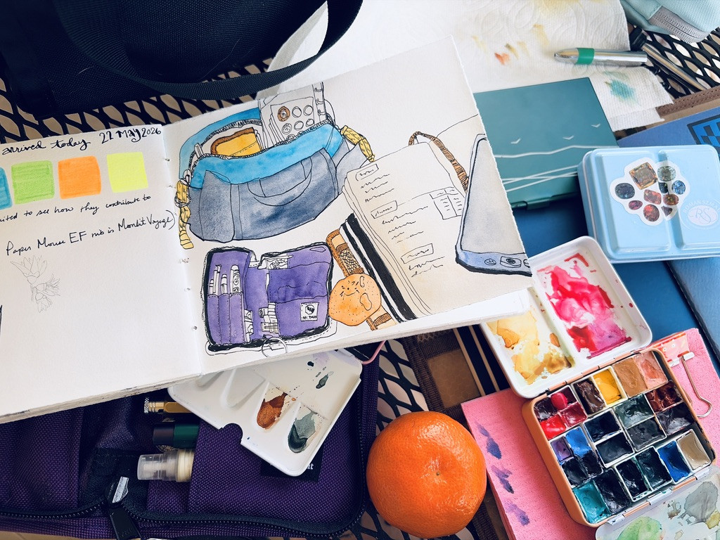 A tabletop outdoors, covered in an art journal entry of a watercolor-painted bag, supplies case, journal, and an orange, and then all those supplies laid out on the table in real life.