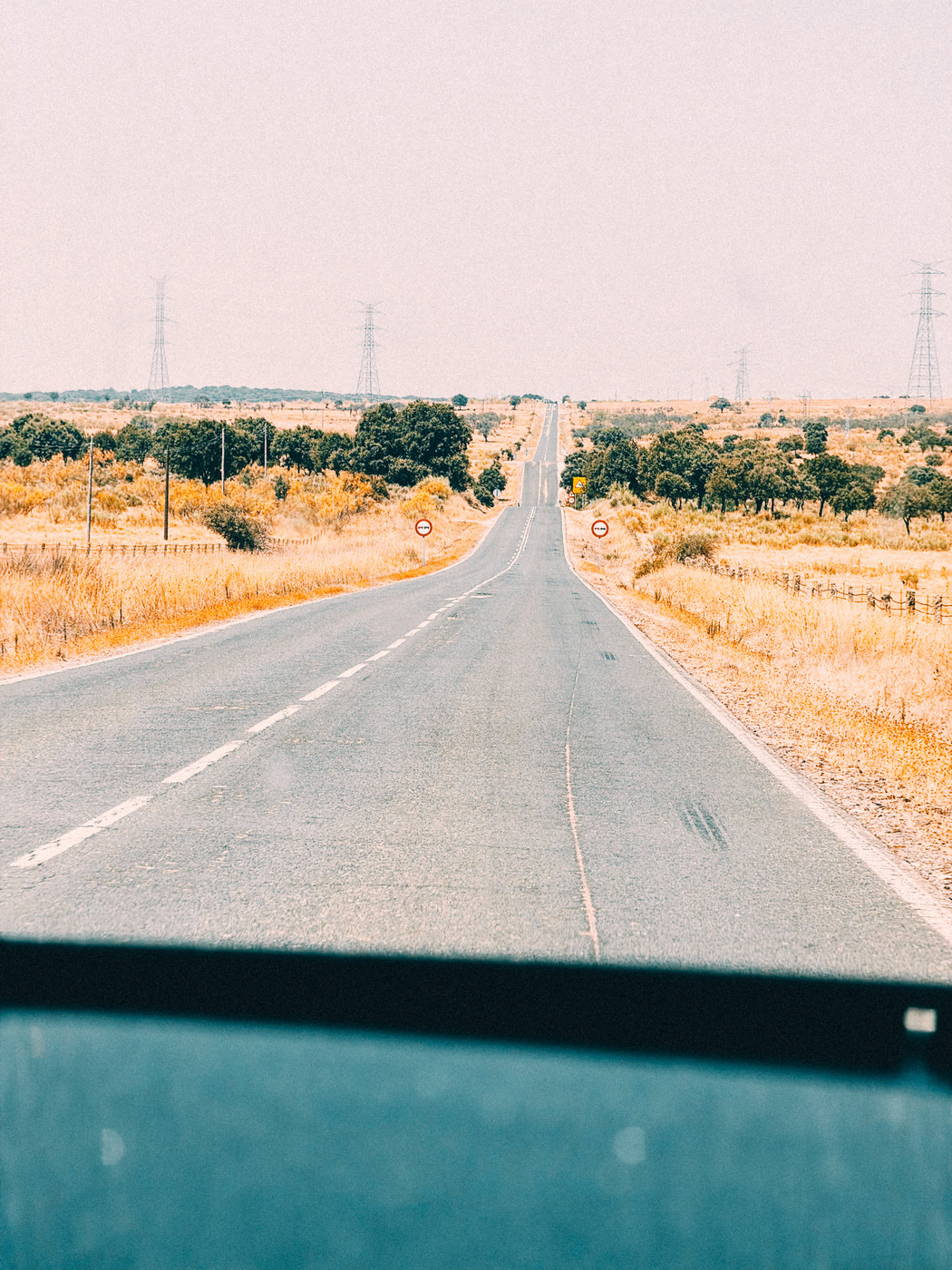 Long, empty rural road stretching into the distance with dry yellow grass on both sides, scattered trees, and power lines under a hazy sky.