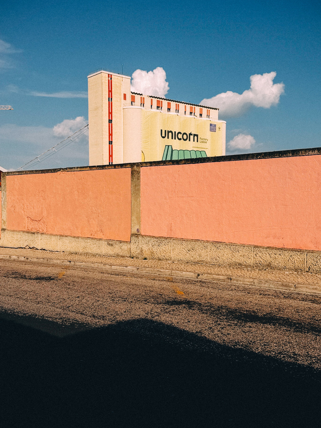 A tall building with a sign reading “Unicorn Factory Lisboa” rises behind a peach-colored wall under a bright blue sky with scattered clouds.