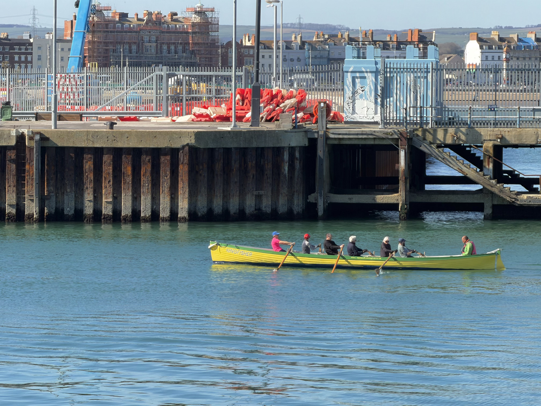 A group rowing a long yellow boat near a pier structure, with calm blue water and buildings lining the shore in the background.