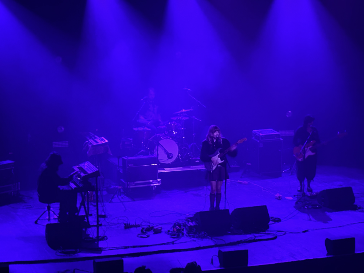 A band performs on stage under deep blue lighting. The group includes a keyboardist, drummer, lead singer with an electric guitar, and a bassist. Amplifiers, microphones, and cables are visible on the floor, and beams of stage light illuminate the musicians through a smoky atmosphere.