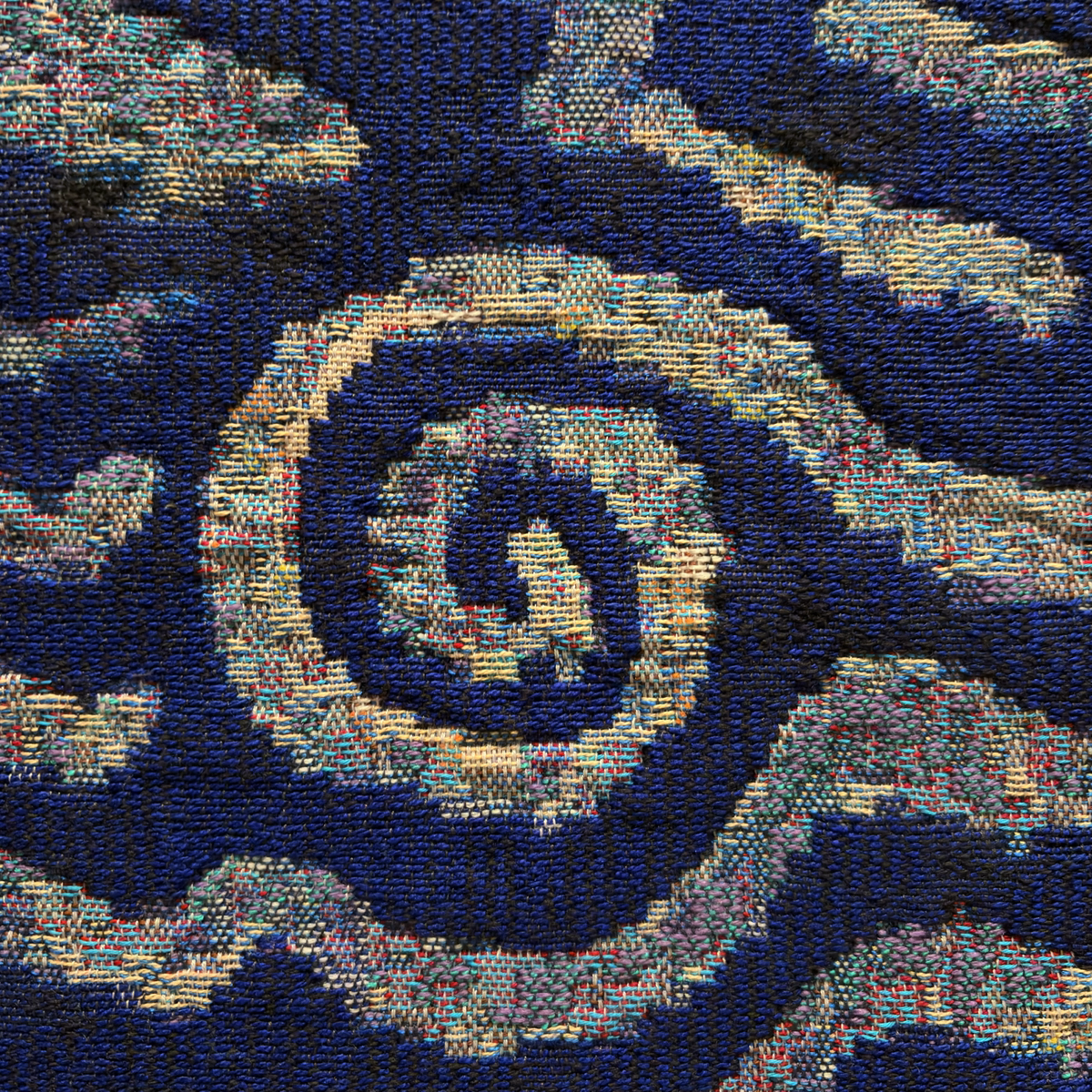 A close-up of a woven textile featuring a spiral pattern in shades of yellow, blue, and multicoloured threads on a dark blue background. The texture of the fabric and individual stitches are clearly visible.