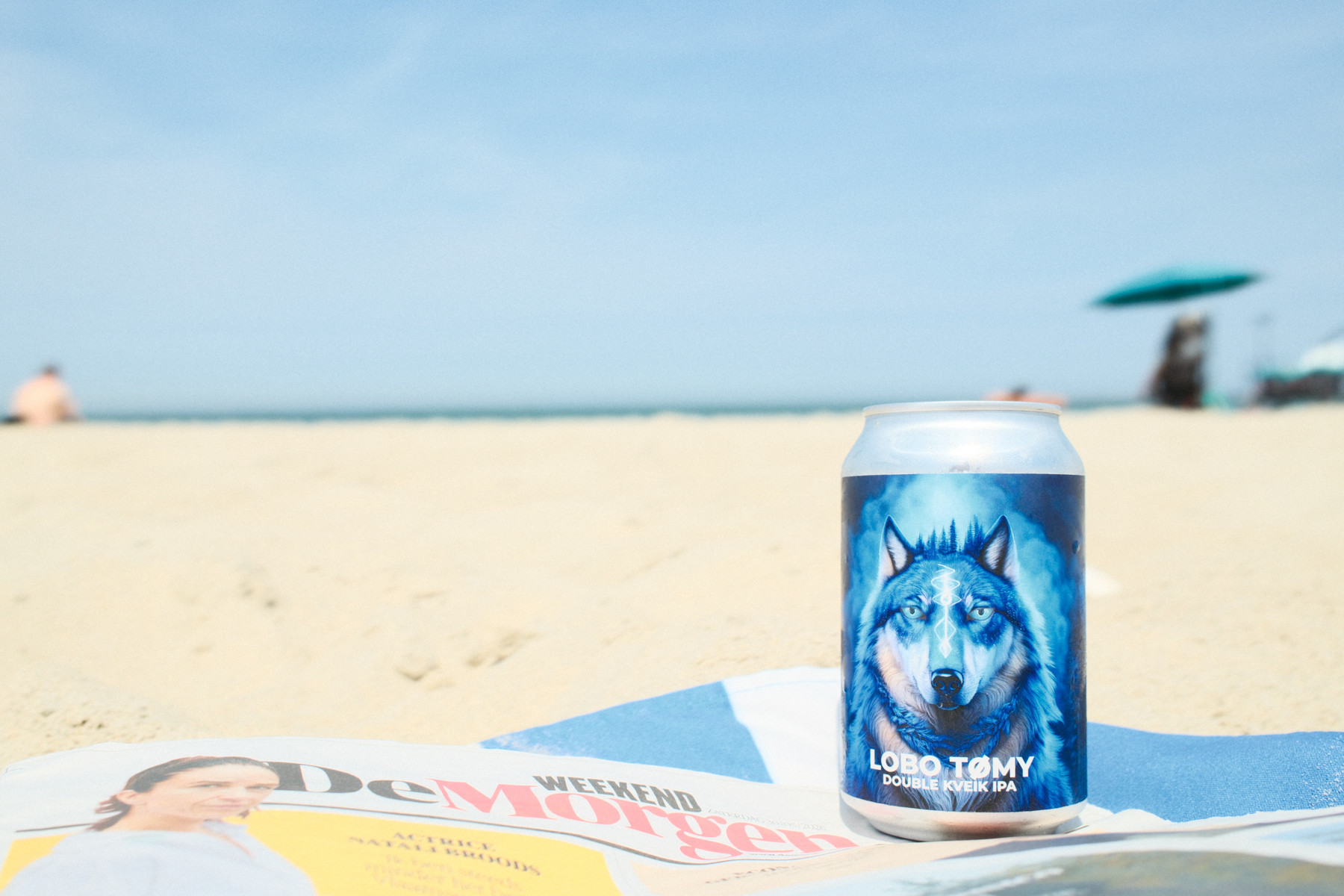 A beer can with a blue wolf illustration sits on a blue-and-white striped towel on a sunny beach. In the blurred background, the ocean, a turquoise beach umbrella, and a person are visible, with a magazine in the foreground.