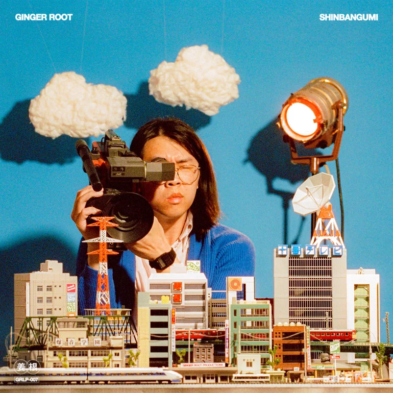 The album cover for Shinbangumi: A person with shoulder-length dark hair and glasses peers through a vintage video camera while positioned behind an intricate miniature model of a Japanese city, complete with tiny buildings, a red transmission tower, a satellite dish, and a commuter train. Fluffy white clouds hang from above on a bright blue backdrop, and a large studio light glows on the right side, casting dramatic shadows. The scene evokes a retro Japanese television studio aesthetic. Text reads "GINGER ROOT" in the top left corner and "SHINBANGUMI" in the top right, with "GRLP-007" and Japanese characters in the bottom left.