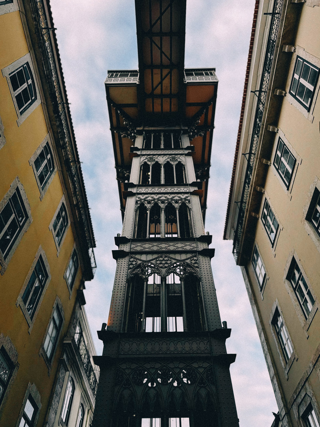 Looking up at the Santa Justa Lift in Lisbon, Portugal, with its ornate iron structure framed by two yellow buildings against a cloudy sky.