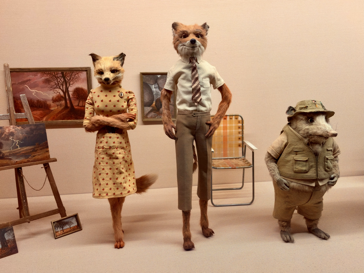 Display of three anthropomorphic animal figures in a gallery setting: a fox woman in a patterned dress with arms folded, a taller fox man in a shirt and tie, and a smaller opossum in a safari-style outfit, with framed paintings, an easel and a folding chair behind them.