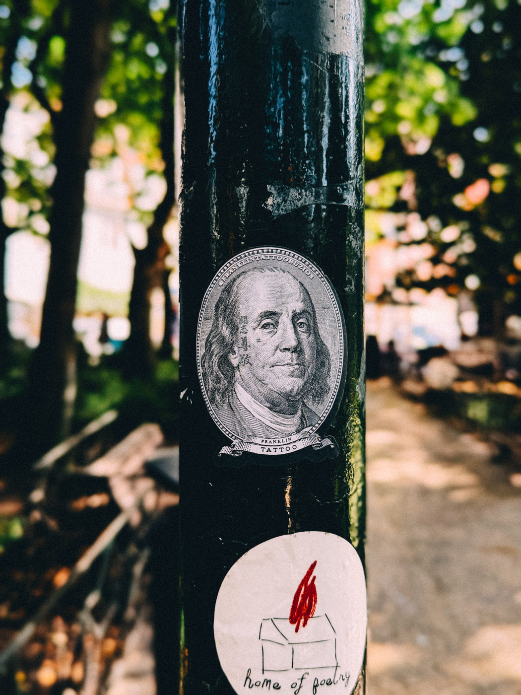 A black pole in a park with two stickers, one showing Benjamin Franklin’s face labeled “Franklin Tattoo” and another depicting a small house with a red flame and the words “home of poetry.”