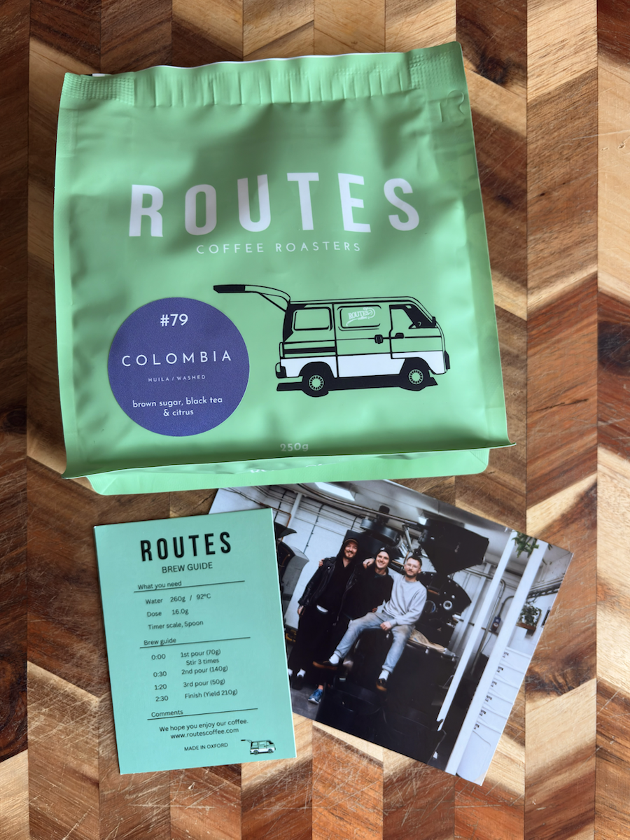 Green bag of Routes Coffee Roasters Colombia #79 coffee, 250g, on a wooden surface, with tasting notes ‘brown sugar, black tea & citrus’ and a small van illustration; beside it are a Routes brew guide card and a photo of three people in a coffee roastery.