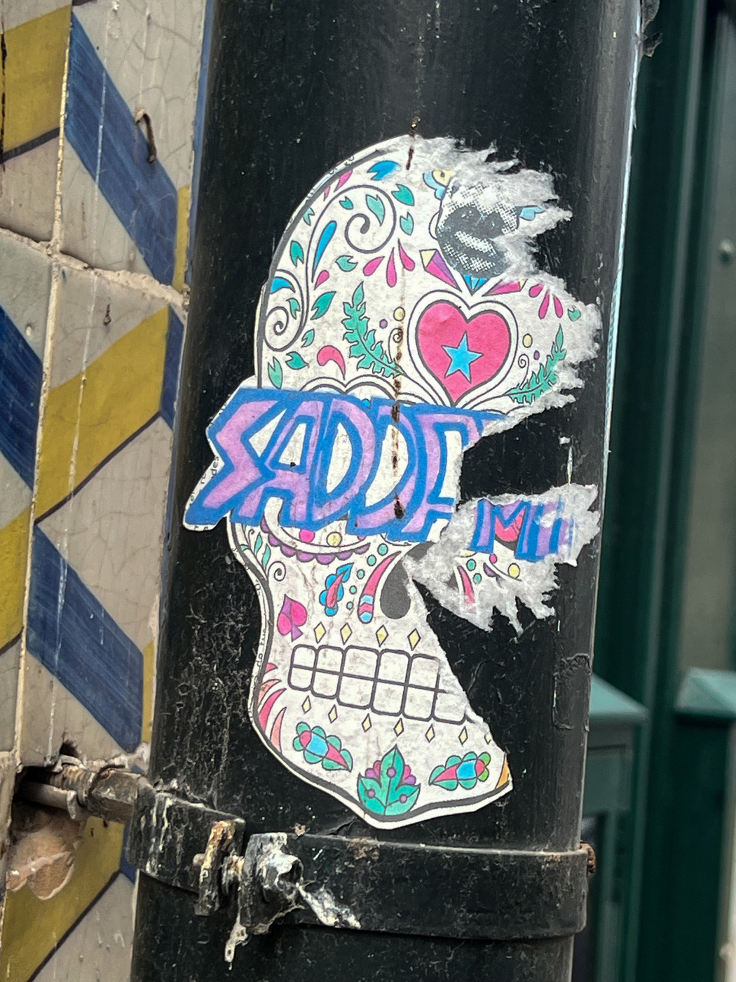 Colorful skull-shaped street art sticker with floral and heart designs, partially torn, on a black pole next to tiled wall with blue and yellow chevrons.