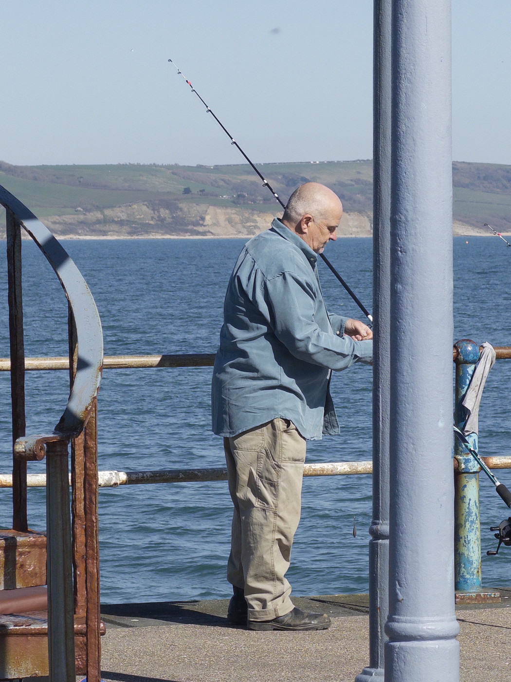 Older man standing on a pier, preparing fishing gear beside a metal post, with calm sea behind him.