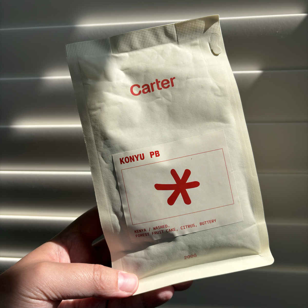 A hand holds a matte white 200 g bag of Carter Coffee’s ‘KONYU PB’ Kenyan washed coffee in front of sunlit window blinds. The minimalist packaging features red branding and a large red asterisk-like symbol, with tasting notes reading ‘forest fruit, fruit cake, citrus, buttery’.
