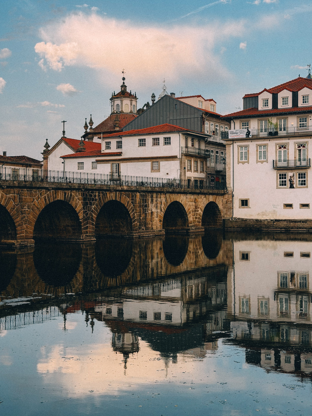 Historic stone bridge with multiple arches over a calm river, reflecting traditional white buildings with red-tiled roofs and a church tower under a partly cloudy sky.