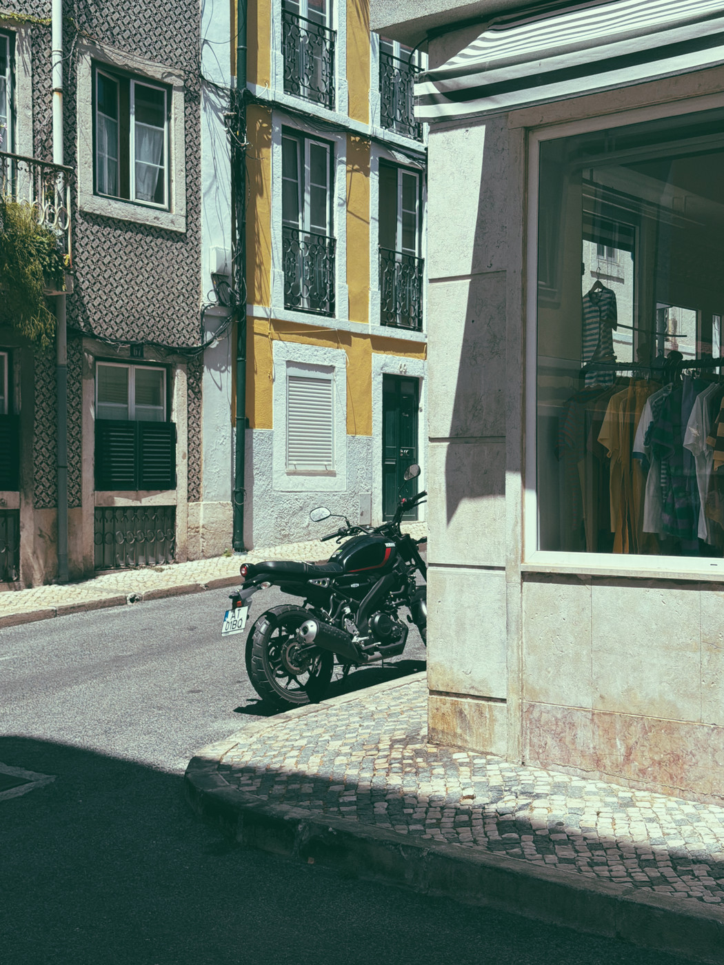 A black motorcycle is parked on a narrow cobblestone street beside a corner clothing shop with a glass window, surrounded by colorful old buildings with balconies and shutters.