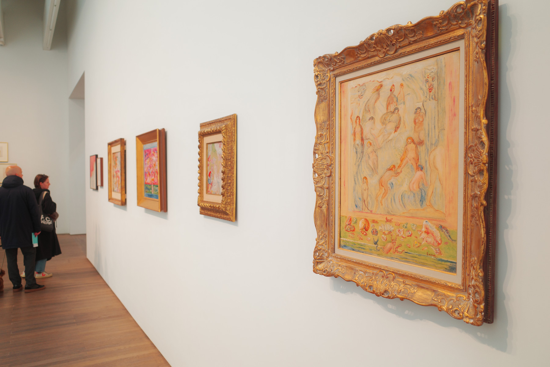 Interior of an art gallery with a row of framed paintings along a pale wall. The closest piece is a large, ornate gold frame housing a pastel, impressionistic scene with numerous small figures. In the background, two visitors in dark coats examine the art, and a wooden floor is visible.