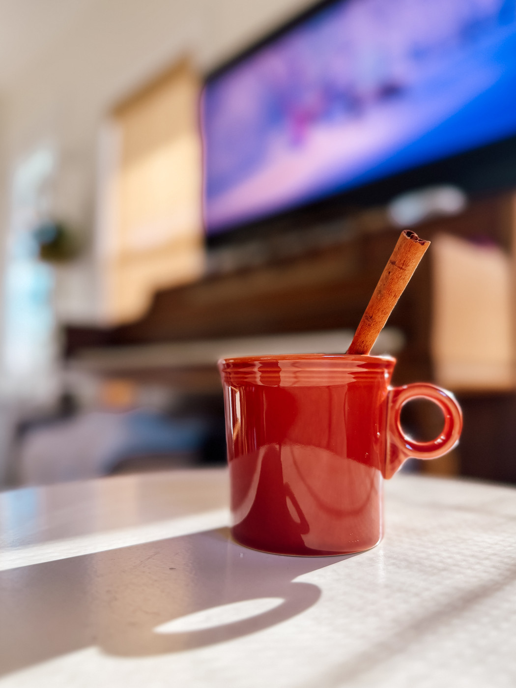 A burnt orange mug with a cinnamon stick sits on a white table in a warmly lit room, with a piano and television in the blurred background.