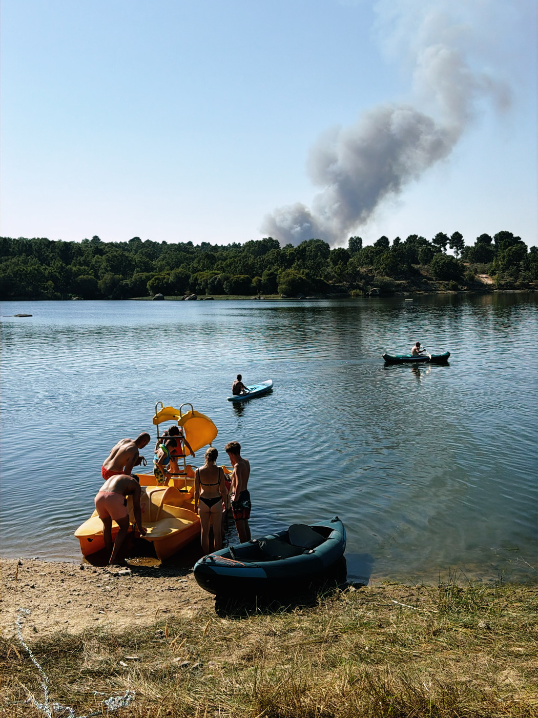 People enjoying boating and swimming at a lake shore with a yellow pedal boat and inflatable kayak, while smoke from a distant forest fire rises into the clear blue sky.