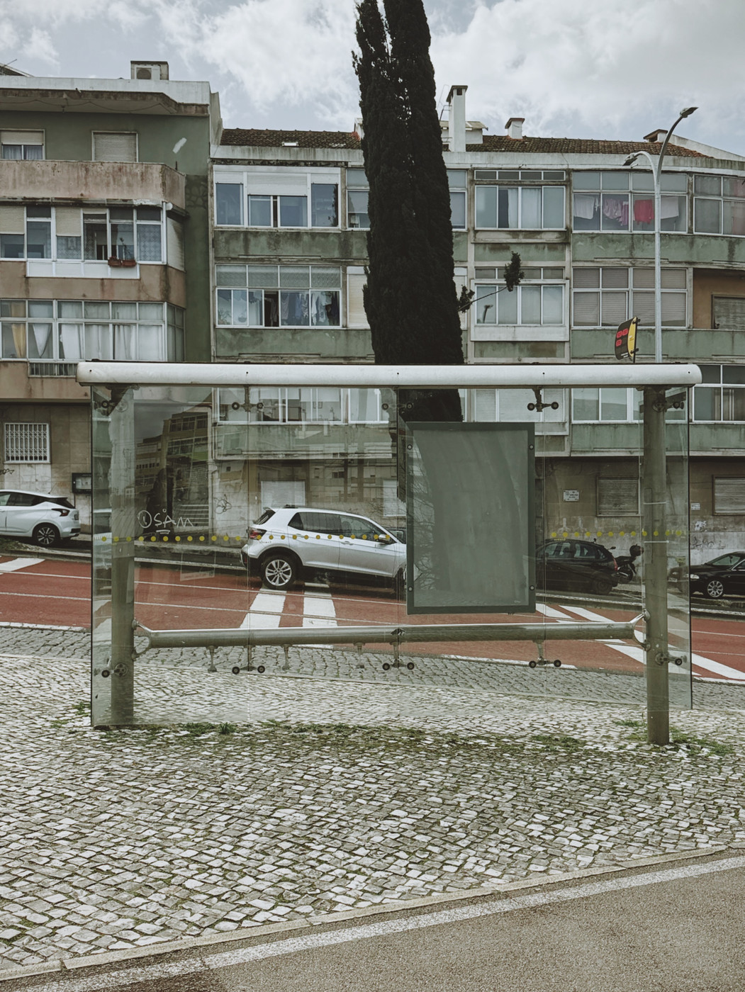 Empty glass bus shelter on a cobblestone sidewalk with apartment buildings and parked cars in the background under a cloudy sky.