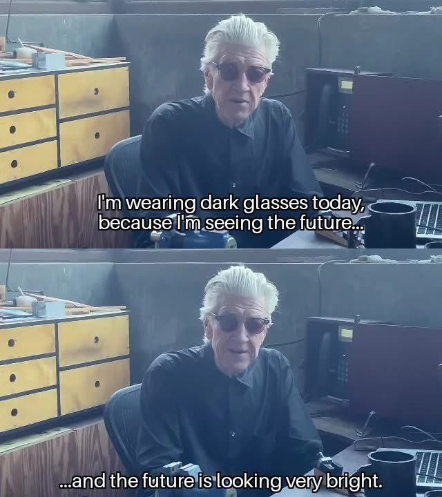 A two-panel vertical collage featuring filmmaker David Lynch in a workshop setting. Top Panel: Lynch is seated at a desk, wearing a black button-up shirt and dark, round sunglasses. He has his signature voluminous, swept-back white hair. He is looking toward the camera with a neutral expression. White subtitle text at the bottom reads: “I’m wearing dark glasses today, because I’m seeing the future...” Bottom Panel: A nearly identical shot of Lynch, though his expression has shifted into a slight, knowing smile. The subtitle text concludes: ”...and the future is looking very bright.” Background: The setting is a cluttered, industrial-style workspace with wooden drawers, a desk lamp, and various tools, creating a utilitarian and atmospheric environment.