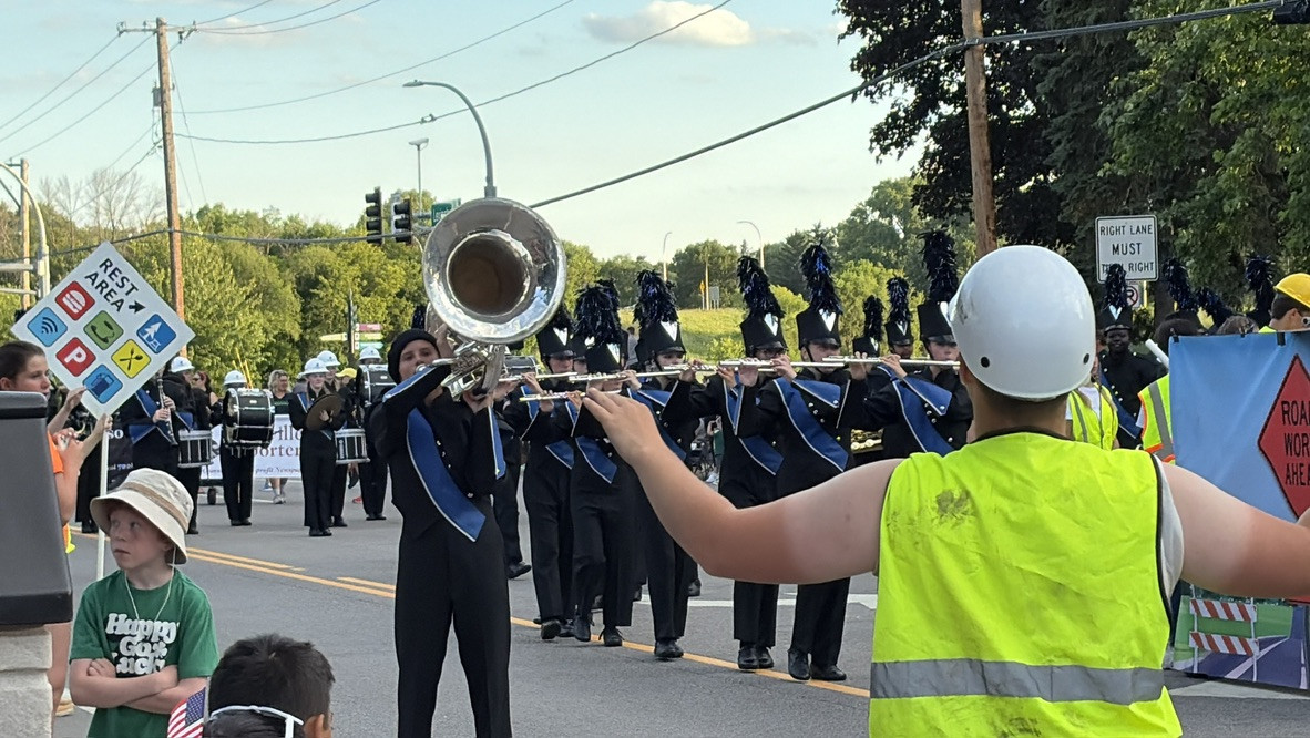 Marching band members perform in a street parade while a construction worker in a hard hat and high-visibility vest stands in the foreground directing them.