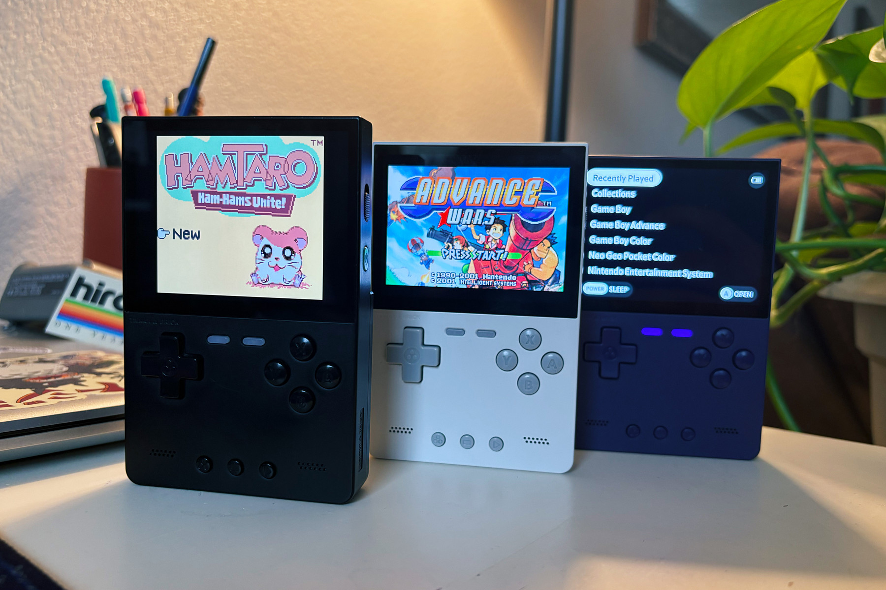 Three handheld gaming devices on a desk, showing retro games on their screens, with a plant and office items in the background.