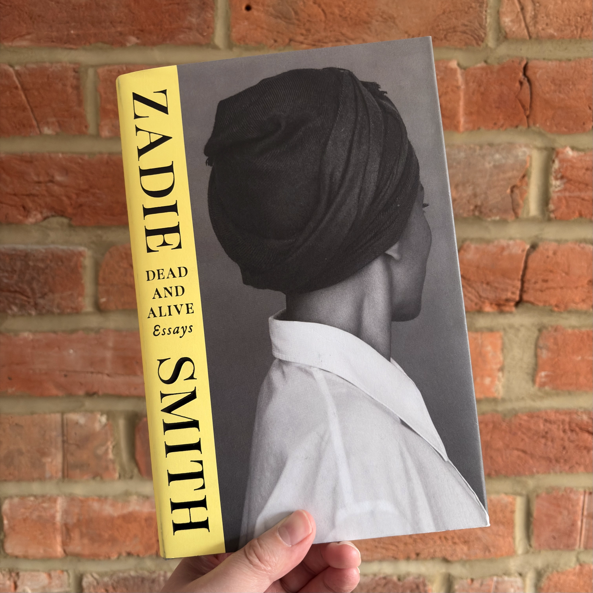 Book cover of ‘Dead and Alive: Essays’ by Zadie Smith, featuring a black and white profile photograph of a woman in a headwrap, with a yellow spine bearing the author’s name, held up against a red brick wall.