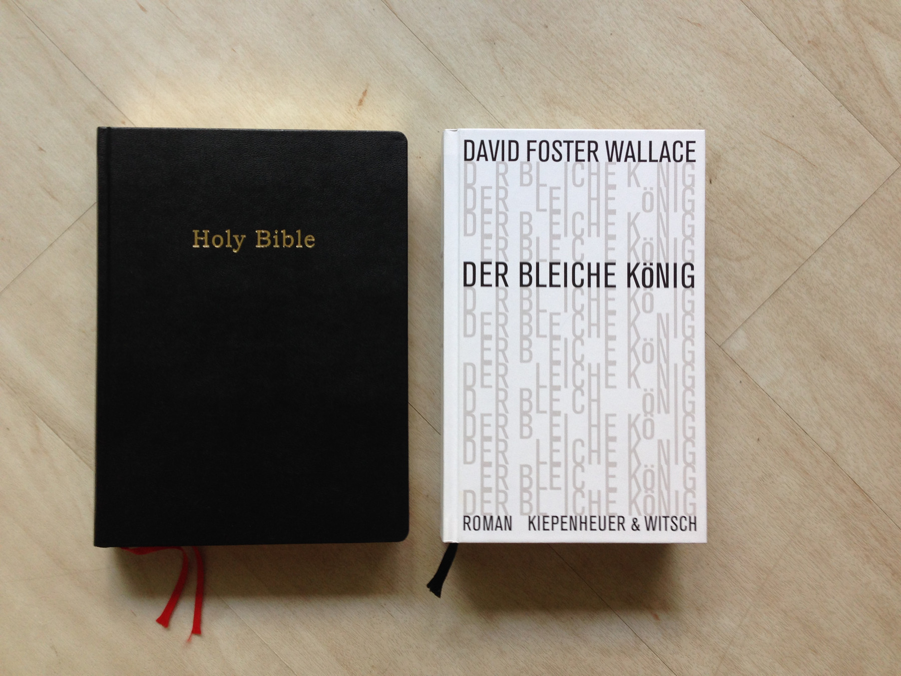 Auf einem hellen, glatten Holzboden liegen zwei dicke Bücher nebeneinander. Auf der linken Seite befindet sich eine englischsprachige Bibel mit einem schlichten, schwarzen Einband und dem in Gold geprägten Titel „Holy Bible“ in der Mitte. Unten schauen zwei rote Lesebändchen heraus. Rechts daneben liegt die deutsche Ausgabe des Romans „Der bleiche König“ von David Foster Wallace mit dem bekannten weißen, typografischen Cover von Kiepenheuer & Witsch. Auch hier ist unten ein Lesebändchen zu sehen, dieses ist schwarz.