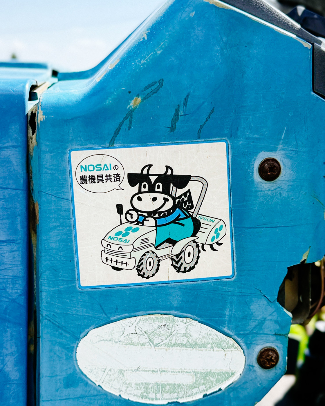 Sticker of a cartoon cow driving a small tractor with Japanese text and the word “NOSAI” on a blue, weathered machine surface.