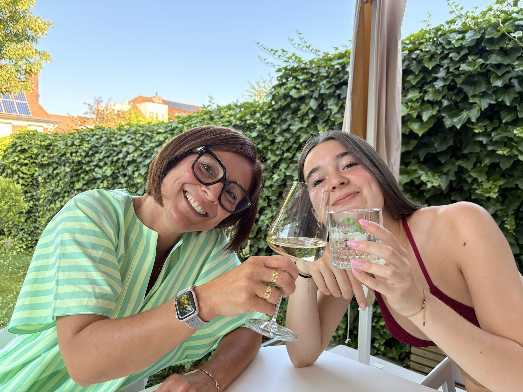 Two women sit at a white outdoor table in a sunny garden, clinking wine glasses and smiling at the camera. On the left, a woman with short brown hair and black-framed glasses wears a green and white striped shirt; on the right, a woman with long hair in a burgundy top wears a delicate necklace. A lush ivy-covered wall and a bright blue sky fill the background.