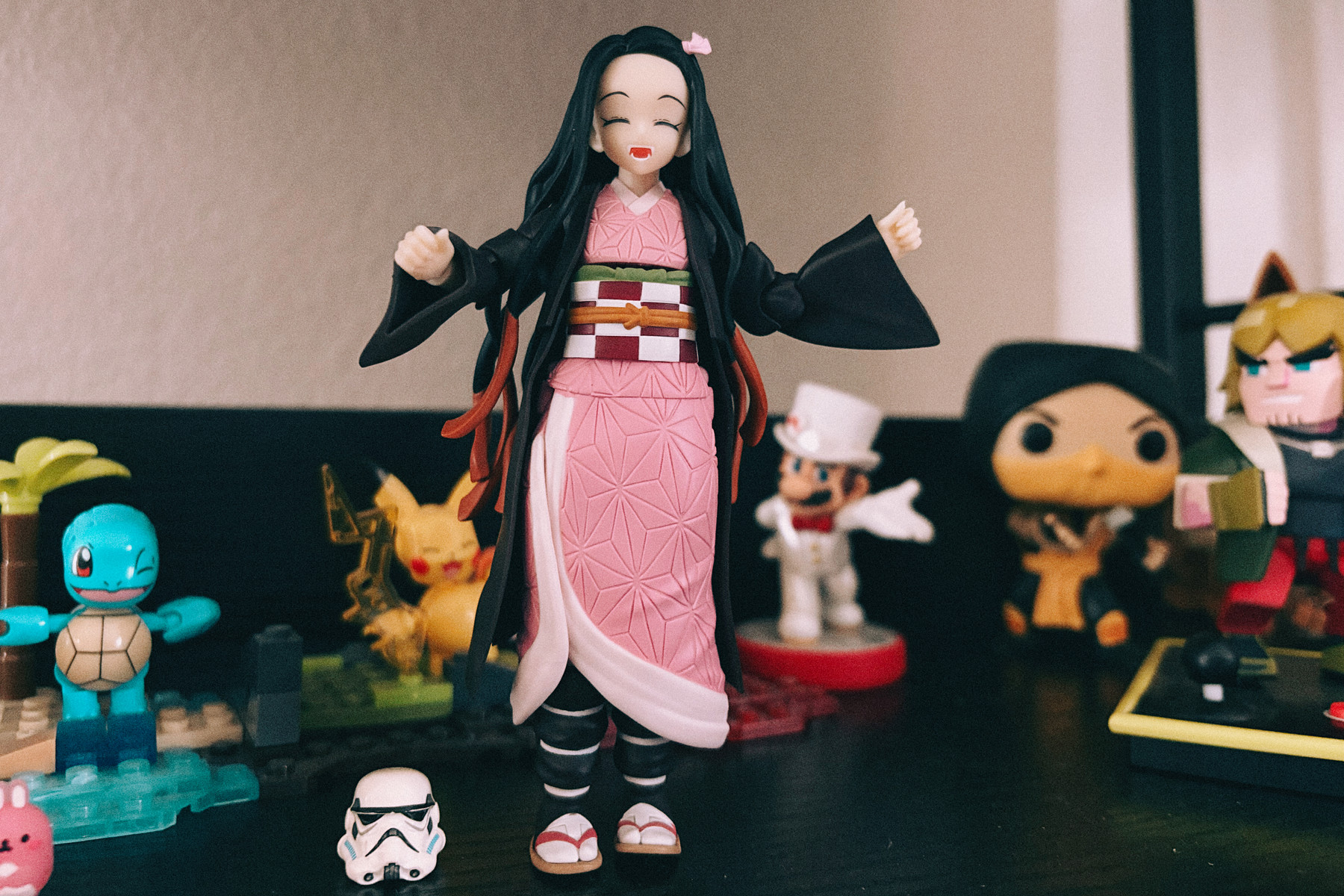 A smiling anime figurine in a pink kimono with long black hair stands centered on a shelf, arms slightly raised. Behind it are various collectible figures, including a Squirtle and Pikachu on the left and other stylized characters on the right, with a small white helmet placed in front.